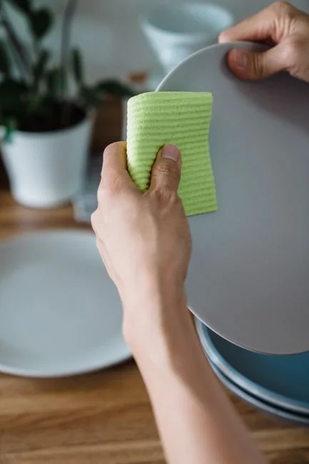 A hand with a feather duster cleaning a wooden desk, with plants and office items all around.